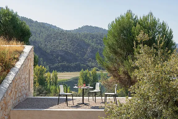 Runder Tisch mit vier Stühlen auf Terrasse mit Blick auf See, Bäume und bewaldete Hügel in mediterraner Landschaft.