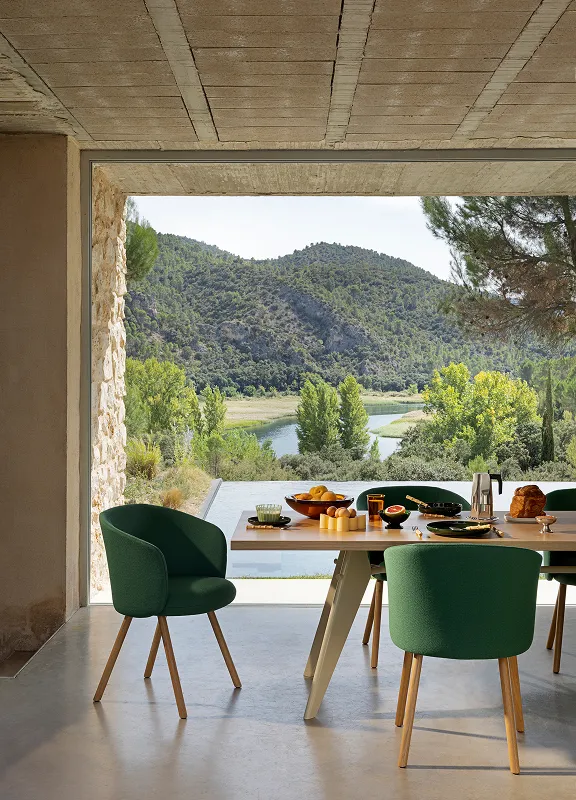 Essbereich mit grün gepolsterten Stühlen und Holztisch vor großer Fensterfront mit Blick auf See und bewaldete Hügel.